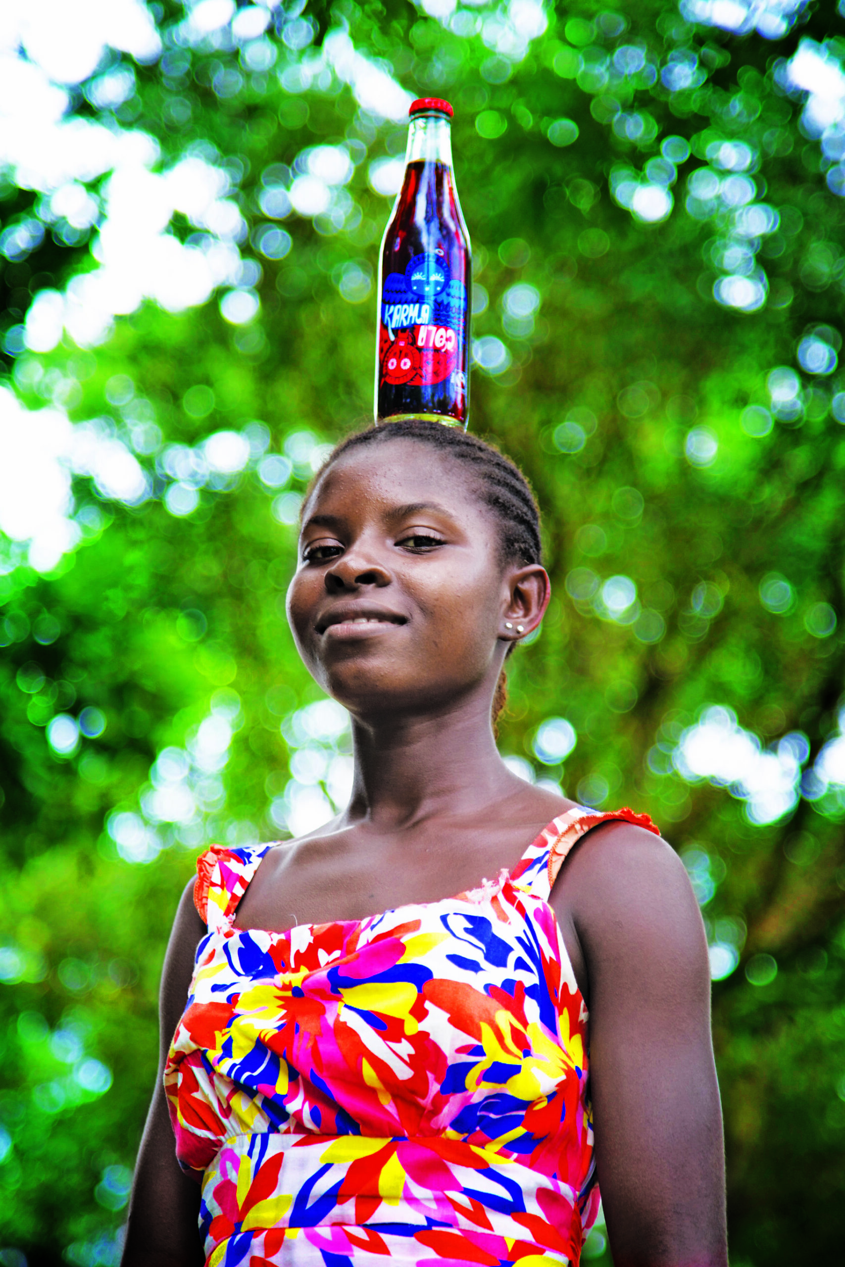 Image of girl standing smiling with bottle of Karma Cola balancing on her head
