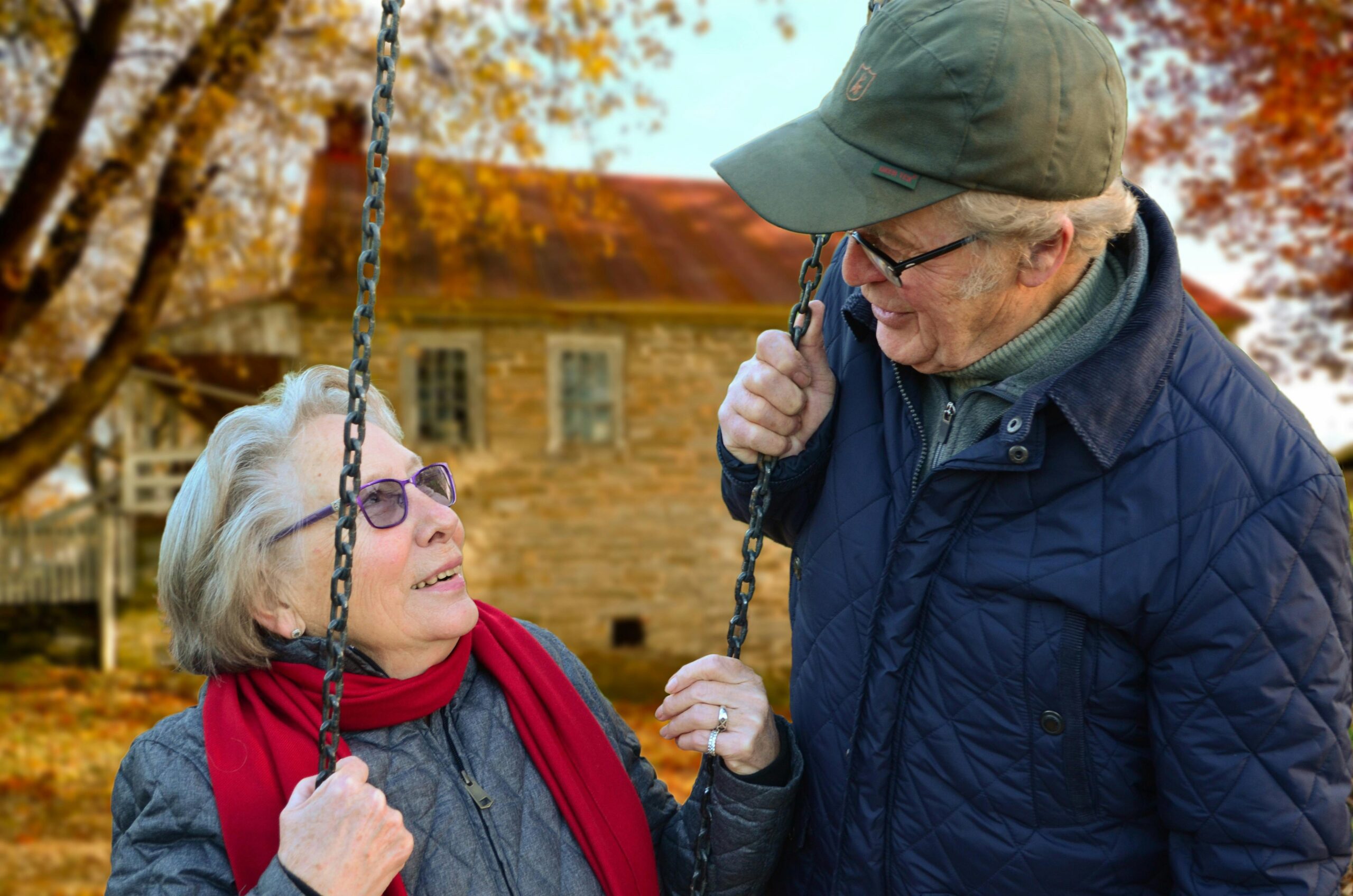 Image of two older people. A woman is sitting on a swing in a garden, and a man is looking at her, smiling.
