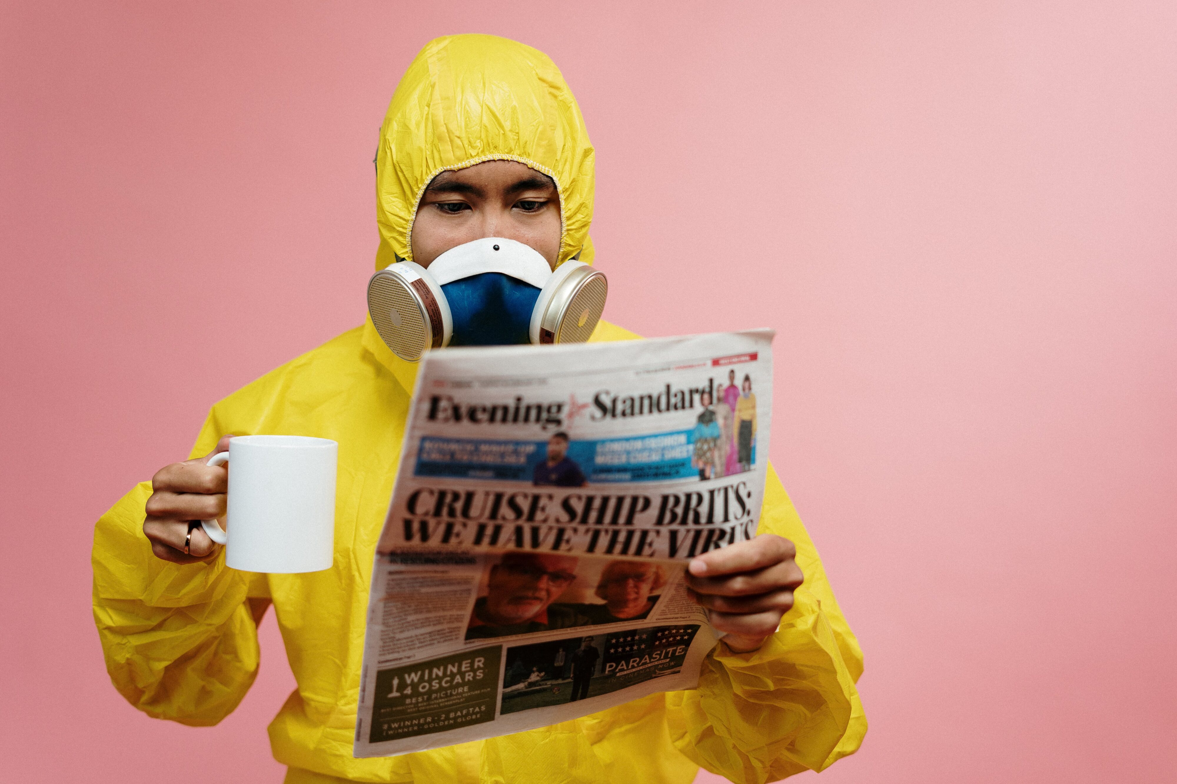 A man in PPE and breathing equipment reads a newspaper in one hand a holds a drink in a mug in the other