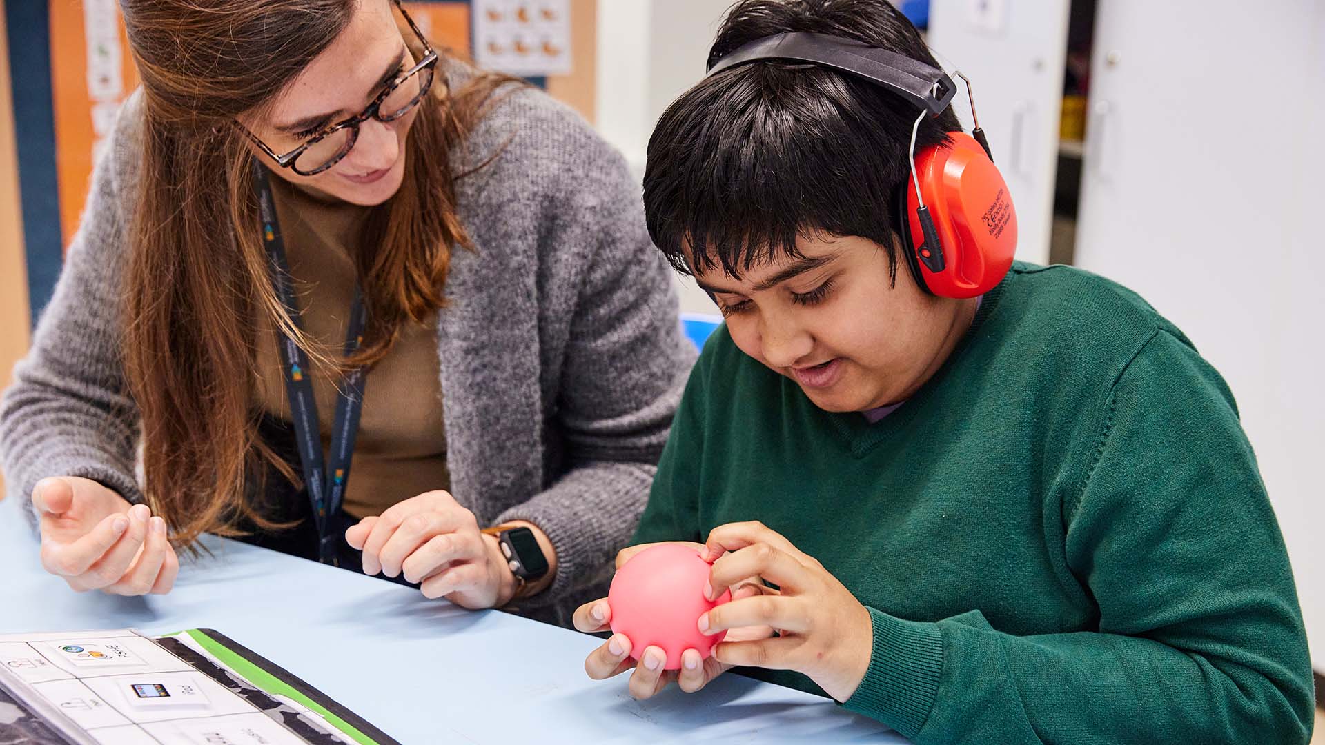 Photo of teacher sitting at a classroom desk with a young autistic person. Both people are smiling and looking down at a small sensory toy the young person is holding.