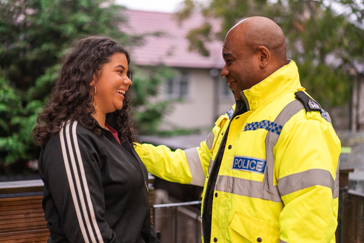 Photo of a police officer wearing high vis jacket standing and smiling. He's looking at his daughter, who's smiling back